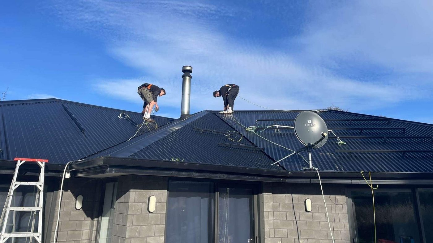 Two Weka Solar installers working on a dark metal roof with a ladder against the eave
