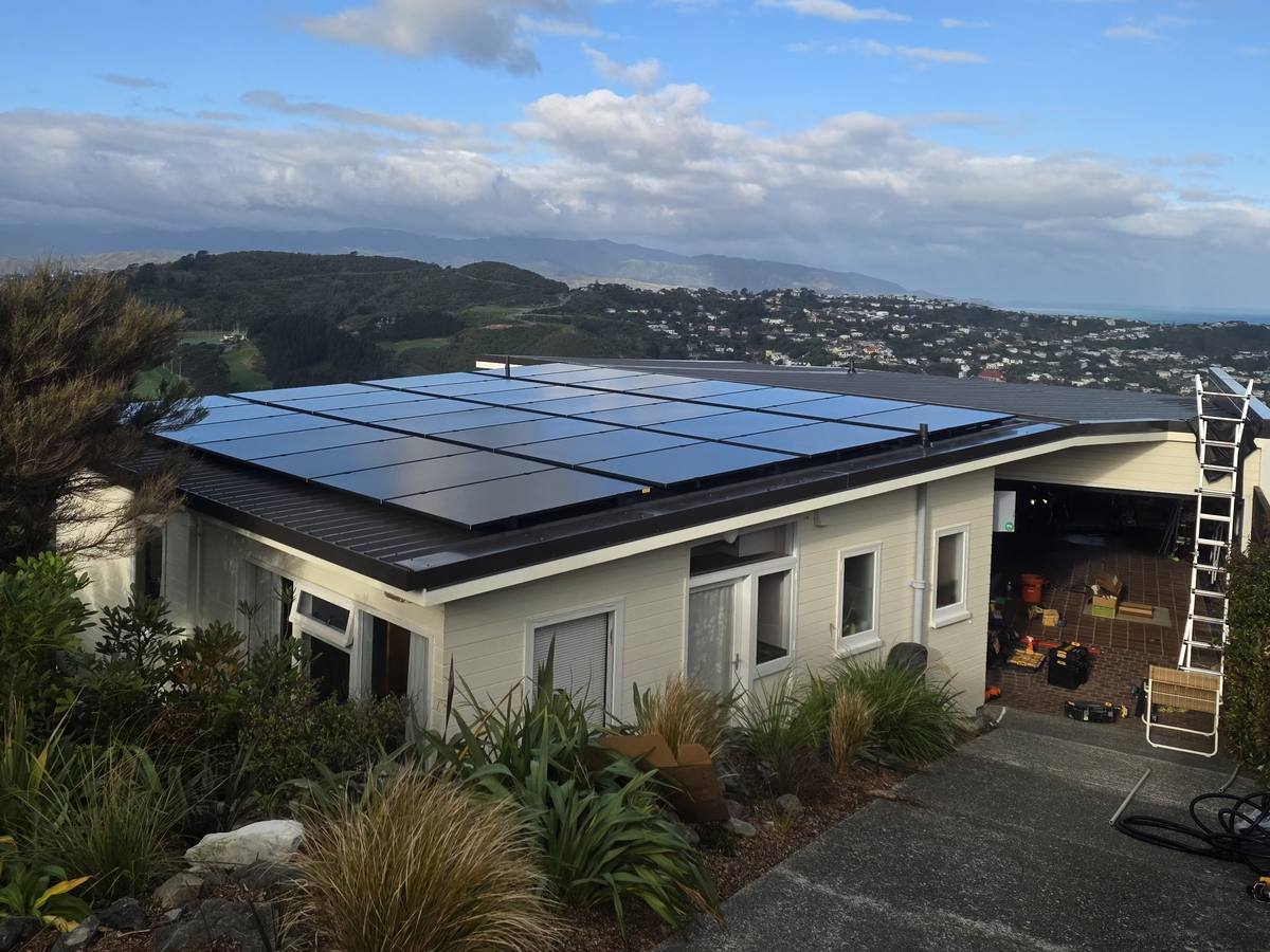 All-black solar panels on the roof of a hillside home, with the suburb and the coast below