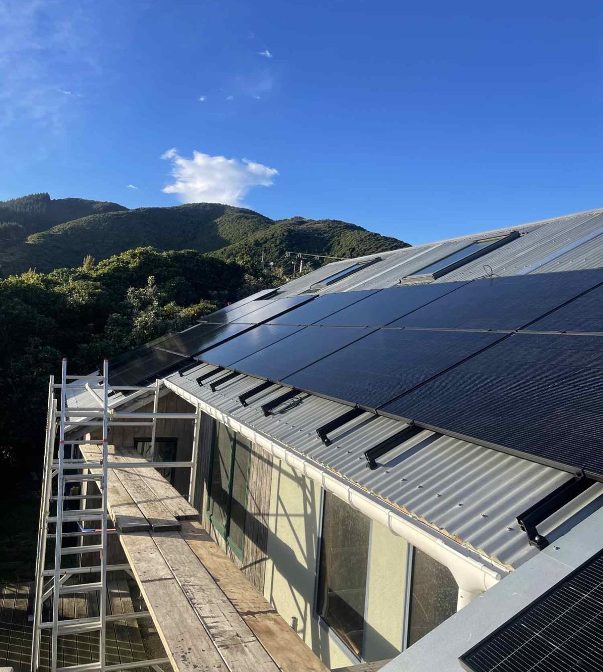 Black solar panels going on to a corrugated metal roof mid-install, scaffolding up and bush-covered hills behind